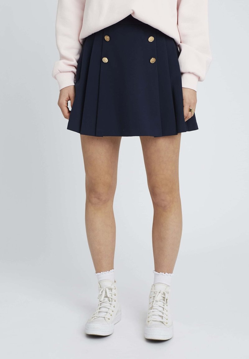 Jupe plissée bleu marine avec double boutons dorés à l'avant, associée à des baskets montantes blanches et des chaussettes côtelées.