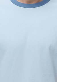 Κοντινή λήψη ενός ανοιχτού γαλάζιου μπλουζάκι με πιο σκούρο μπλε στρογγυλό λαιμόκοψη στο πάνω μέρος του στήθους και του λαιμού ενός ατόμου.