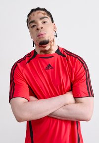 Red Adidas athletic shirt with black accents and stripes. Short sleeves, crew neck design. Smooth texture, fitted silhouette.