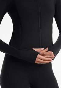 Giacca atletica aderente nera con colletto alto, chiusura a zip, maniche lunghe e aperture per il pollice. Texture liscia e senza modelli o loghi visibili.