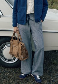 Veste en polaire bleue, pantalon large gris clair, baskets bleu marine et sac à main en daim marron. Tissu texturé avec poches visibles et détails métalliques.