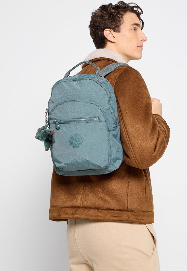 Mochila azul claro con una superficie texturizada, bolsillo frontal con cremallera y un logo circular. Cuenta con un llavero de peluche como accesorio.