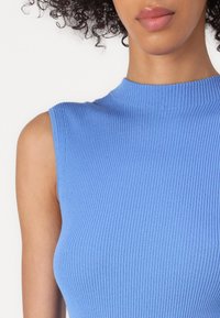 Woman with curly hair wearing a sleeveless, ribbed light blue top, shown from shoulders to lips against a white background.