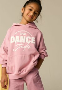 Różowa bluza z kapturem z białym napisem "DANCE Studio" i kieszenią kangurką, zestawiona z różowymi spodniami w paski. Swobodny, luźny krój.