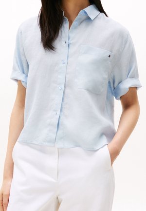 Kvinde iført lys blå kortærmet skjorte med knapper og brystlomme samt hvide bukser, med den ene hånd i lommen, foran en ensfarvet baggrund.