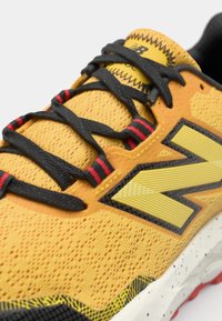 Gul atletisk sko med en teksturert mesh-overdel, svarte snørebånd og røde detaljer. Har et karakteristisk logo og en polstret såle.