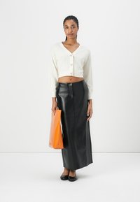 Un cardigan cropped blanc en tricot avec des manches ballon, associé à une jupe maxi taille haute en cuir noir. Le mannequin tient un sac tote orange.