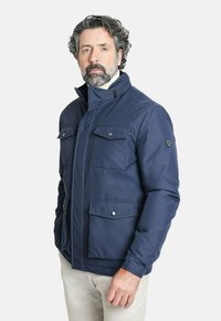 Chaqueta aislante de color azul marino con alto cuello, dos grandes bolsillos frontales y botones a presión. Textura suave y ajuste entallado.