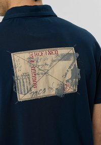 Mornarska polo majica s teksturiranim materijalom i velikim otiskom razglednice na leđima s izblijedjelim grafičkim dizajnom i crvenim naglascima.