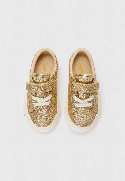Um par de ténis para bebés brilhantes em dourado com solas brancas, tiras de velcro e atacadores em creme sobre um fundo branco liso.