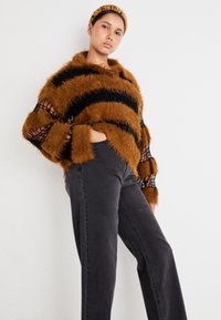 Moteris vilkinti pūkuotą rudos ir juodos juostuotos megztinio su tinklinėmis rankovėmis, juodas aukšto juosmens džinsus ir atitinkantį rudą šalį.