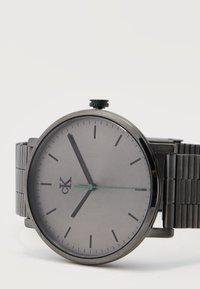 Montre-bracelet grise minimaliste Calvin Klein avec aiguilles des heures et des minutes noires, trotteuse verte et bracelet à maillons métalliques sur fond blanc.
