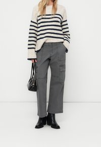 Pull en maille épaisse avec des rayures crème et bleu marine, associé à un pantalon cargo gris avec des poches, et des bottines à talons noires. Petit sac à main noir.