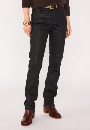 Pantaloni in pelle neri con vestibilità slim, caratterizzati da una superficie liscia e una leggera lucentezza, abbinati a stivali in pelle marrone con tacco. Dettagli in metallo dorato.