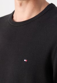 Gros plan sur une personne portant un pull noir à col rond avec un petit logo rectangulaire rouge, blanc et bleu sur la poitrine.