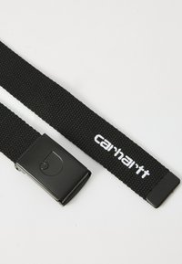 Ceinture tressée noire avec une boucle métallique rectangulaire noire, arborant un logo brodé blanc. Tissu durable, design droit et surface texturée.