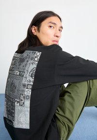 Czarna koszulka z długim rękawem z dużym szarym nadrukiem na plecach, zawierającym różne symbole. Model siedzi w luźnej pozycji, w oliwkowych spodniach.
