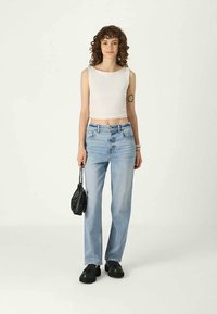 Jovem mulher com cabelo encaracolado, vestindo uma blusa branca sem mangas, jeans de cintura alta azul claro, sapatos pretos, segurando uma pequena bolsa preta.