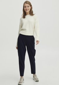 Cream textured sweater with long sleeves paired with dark navy tapered pants and gray sneakers. Focus on simplicity and comfort in design.