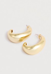 Une paire de boucles d'oreilles créoles dorées, épaisses et courbes, avec des attaches en plastique transparent sur un fond clair.