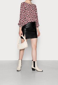 Langärmelige Bluse mit Muster in Burgunderrot und Creme, kombiniert mit einem schwarzen Minirock aus Leder und cremefarbenen Stiefeletten. In der Hand eine geflochtene cremefarbene Handtasche.