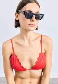 Κόκκινο μπικίνι με στρογγυλεμένες άκρες και λεπτές τιράντες, συνδυασμένο με γωνιακά μαύρα γυαλιά ηλίου, σε ουδέτερο φόντο.