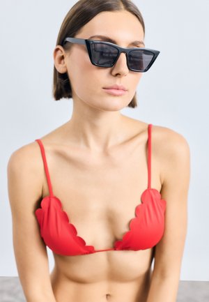 Rød bikini med bølgede kanter og tynde stropper, parret med kantede sorte solbriller, foran en neutral baggrund.