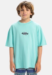 Camiseta de algodón turquesa con cuello redondo y mangas cortas, con un logo gráfico en rosa y negro en el pecho, corte relajado.