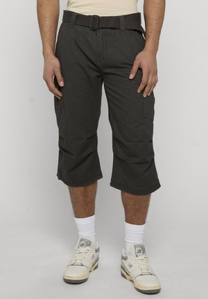 Mand iført sorte cargoshorts, hvide sokker og hvide sneakers, stående mod en ensfarvet lys baggrund.