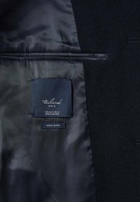 Donker wollen blazer met een binnenvoering van marineblauwe satijn. Labels tonen de merknaam en onderhoudsinstructies, inclusief het label "Wool Blend".