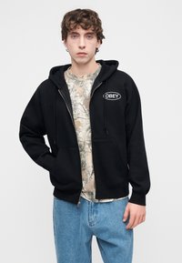 Μαύρο φούτερ με φερμουάρ και λογότυπο στο μπροστινό μέρος, σε συνδυασμό με T-shirt με σχέδιο καμουφλάζ. Το μοντέλο φοράει ανοιχτό μπλε τζιν παντελόνι.