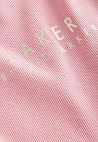 Lichtroze ribstof met verhoogde metallic gouden tekst die "BAKER" spelt, met daaronder kleinere tekst "by TED BAKER".