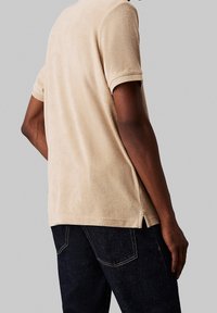 Chemise beige à manches courtes en tissu doux et texturé, avec des fentes sur les côtés, assortie à un jeans en denim foncé. Le matériau est pelucheux et décontracté.