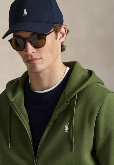 Πράσινο φούτερ με φερμουάρ και λευκό λογότυπο, φορεμένο πάνω από μπλε σκούρο πουλόβερ με στρογγυλή λαιμόκοψη. Συνοδεύεται από μπλε σκουρόχρωμο καπέλο και γυαλιά ηλίου με σκελετό από φολιδωτό σχέδιο.