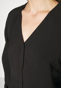 Gros plan d'une personne portant un blazer noir ajusté en V, montrant les détails de l'encolure et du haut du torse avec des coutures visibles.