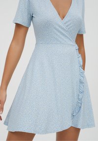 Vestido envolvente azul claro con pequeños patrones florales blancos. Presenta mangas cortas, un escote en V y un detalle de volantes en el lateral. Textura de tela suave.