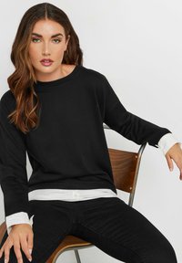 Mustat pitkät hihaiset collegepaidat, joissa on pyöreä kaula-aukko ja valkoiset kontrastihihansuut, yhdistettynä mustiin farkkuihin, istuu puutuolilla.