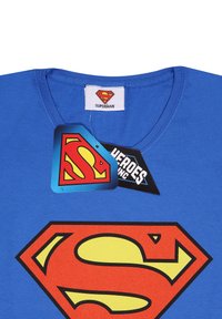 Modrý tričko s výstřihem do V s velkým červeným a žlutým logem Supermana potisknutým na přední straně a s odpovídajícím logem na štítku na límci a připevněnou cenovkou.