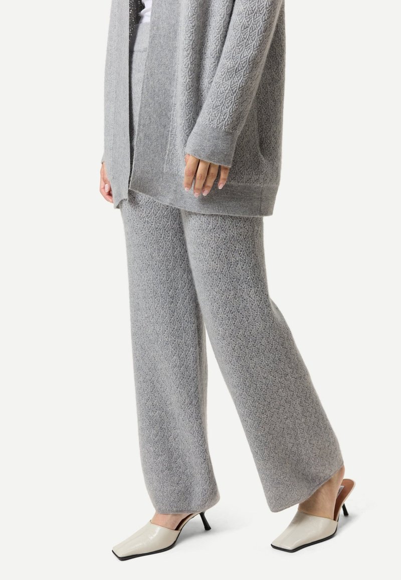 Person trägt graue, strukturiert gestrickte Hosen und passende Strickjacke mit cremefarbenen, quadratischen, hochhackigen Maultieren vor weißem Hintergrund.