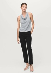 Top grigio con scollo a cowl realizzato in un tessuto morbido, abbinato a pantaloni neri slim. La modella indossa tacchi bianchi con cinturini, in piedi su uno sfondo neutro.