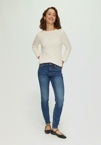 Pull en tricot crème avec un col rond, associé à un jean skinny bleu et des ballerines noires. Le modèle se tient debout, les mains dans les poches, souriant.
