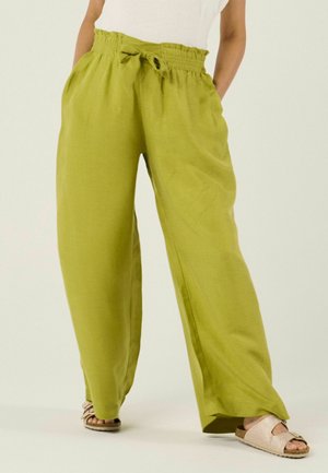 Personne portant un pantalon ample taille haute vert olive avec une ceinture à nouer et des sandales beiges à bout ouvert, debout les mains dans les poches.