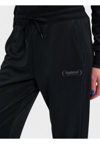 Pantalon de jogging noir avec poches latérales, taille élastiquée avec cordon et logo "hummel sportswear" sur la cuisse supérieure, porté par une personne avec du vernis à ongles bleu.