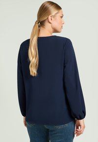 Granatowa bluzka z długimi, zwiewnymi rękawami, okrągłym dekoltem i gładką fakturą. W połączeniu z jasnoniebieskimi dżinsami, widok z tyłu.