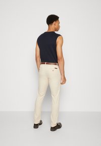 Camiseta sin mangas azul marino, pantalones beige ajustados, cinturón marrón, textura suave y zapatos oscuros. Posicionado de espaldas, mirando hacia la izquierda.