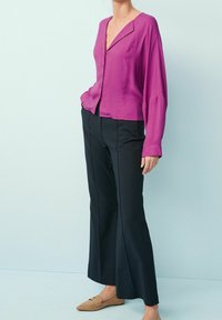 Top button-up di tessuto seta viola con un colletto ampio, abbinato a pantaloni neri a zampa d'elefante. L'outfit è completato da scarpe beige senza lacci.