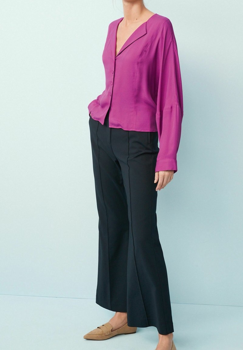 Top button-up di tessuto seta viola con un colletto ampio, abbinato a pantaloni neri a zampa d'elefante. L'outfit è completato da scarpe beige senza lacci.