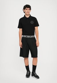 Cămașă polo neagră cu mâneci scurte și un logo circular pe partea stângă a pieptului, asortată cu pantaloni scurți negri, lejeri, cu un tiv marcă.