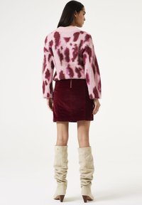 Un pull rose avec des motifs abstraits plus foncés, associé à une jupe en velours côtelé bordeaux et des bottes beiges hauteur genou, flasques et à talons pointus.