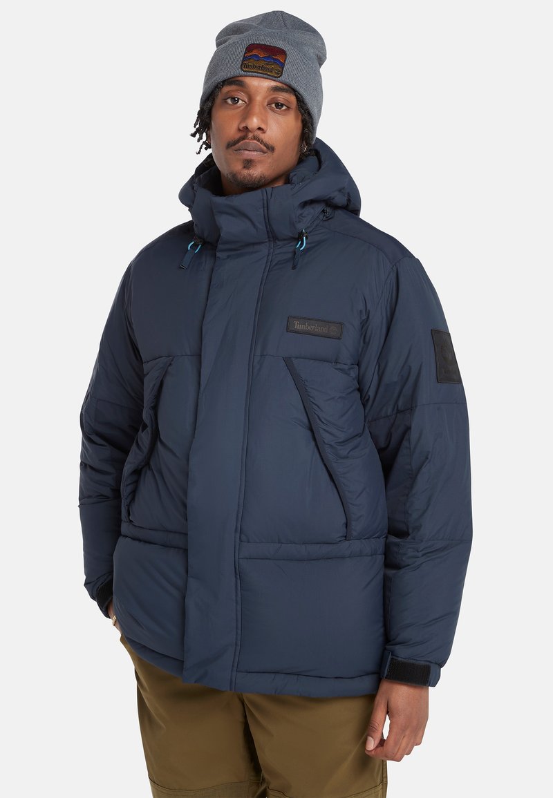 Granatowa kurtka puchowa z wysokim kołnierzem, przednimi kieszeniami i naszywką z logo. Model nosi również szary beret z emblematem.